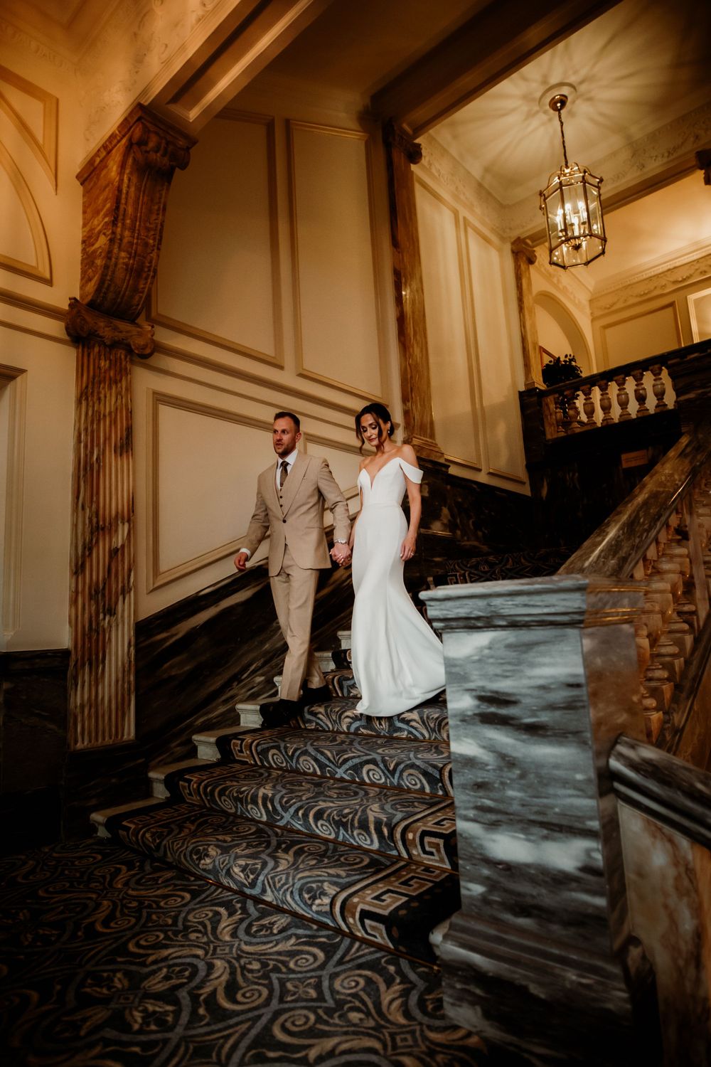 Couple walking down elegant marble staircase with ornate carpet and classical architectural details in grand historic venue.