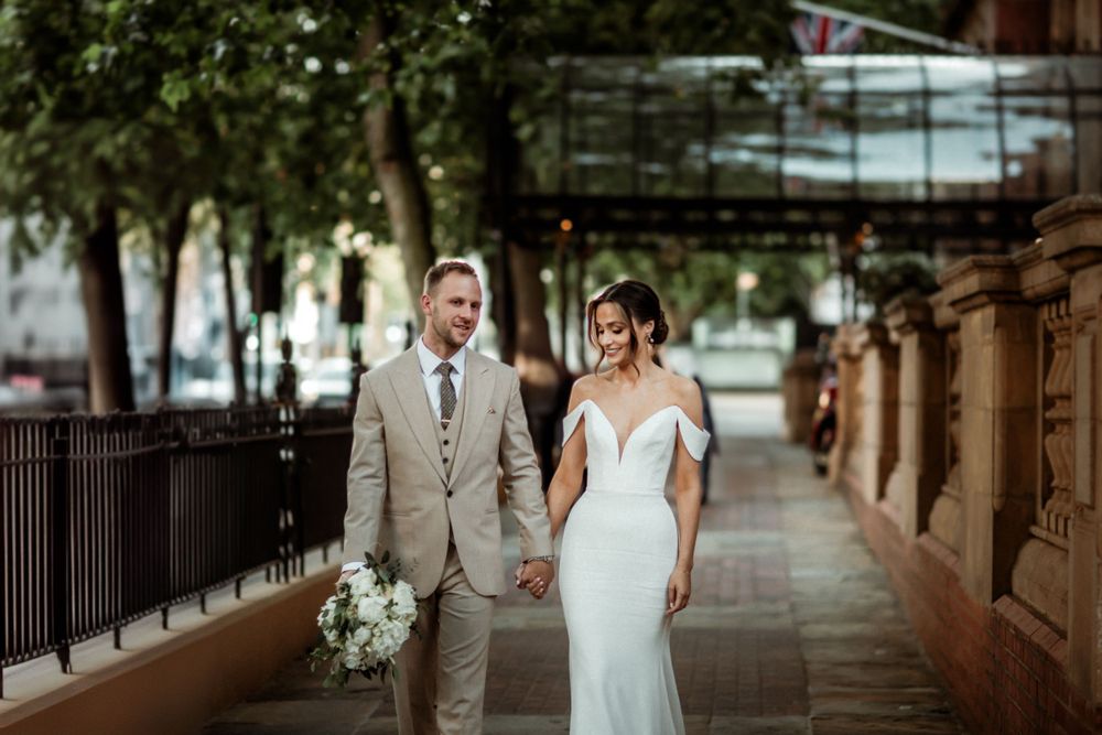 A newlywed couple holding hands and walking together on a city sidewalk lined with trees and iron railings.