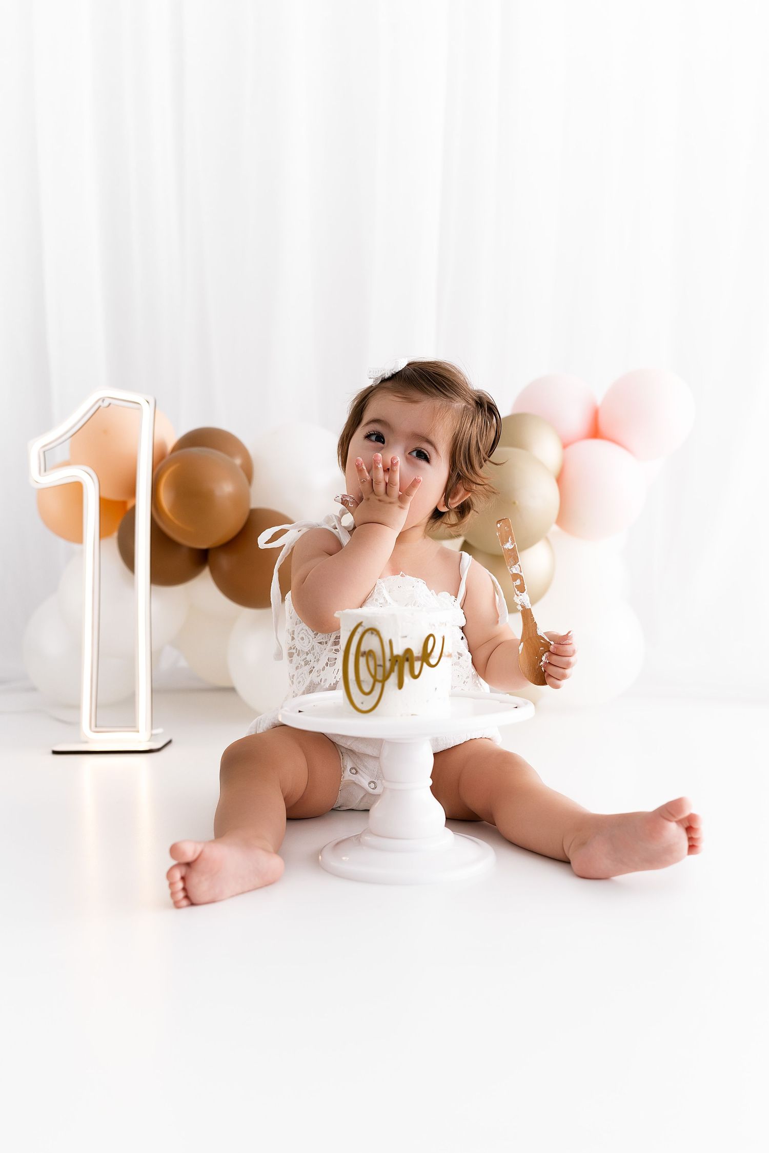 Baby in white outfit sits by first birthday decorations with balloons in background.