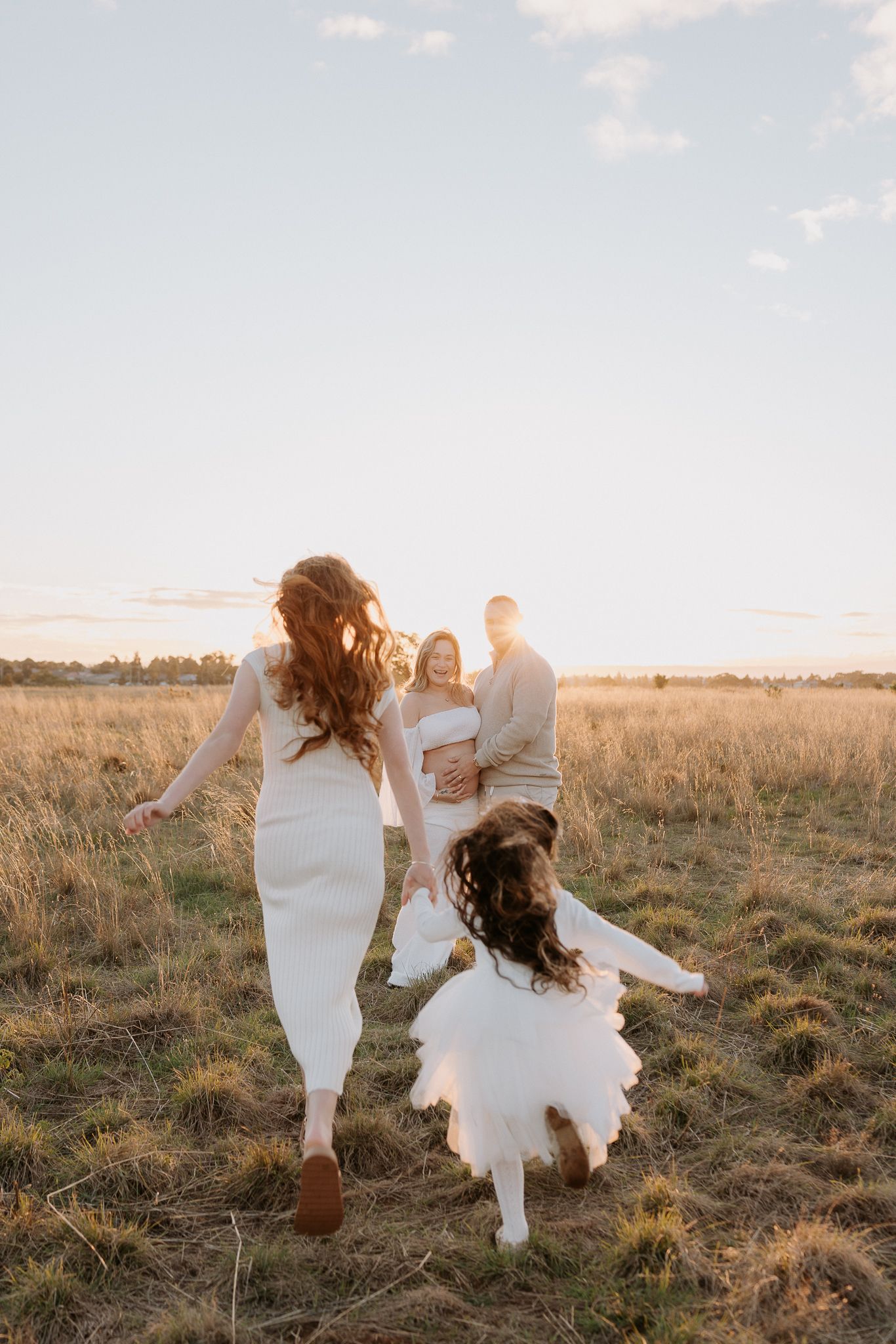 Pregnant woman in white dress poses on beach near rocky cliffs at sunset.