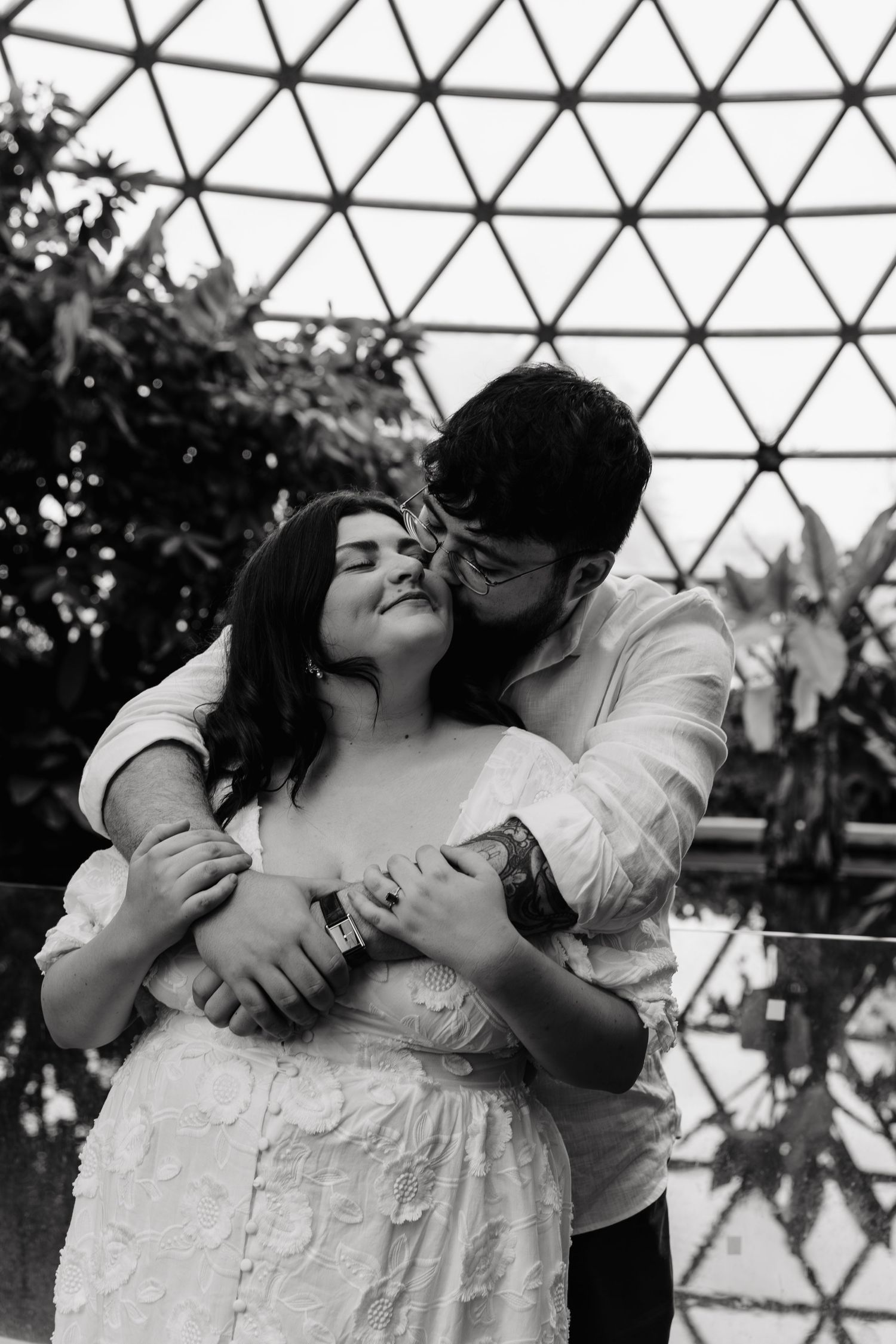 A romantic black and white photo of a couple embracing inside a geodesic dome structure with plants visible in background.
