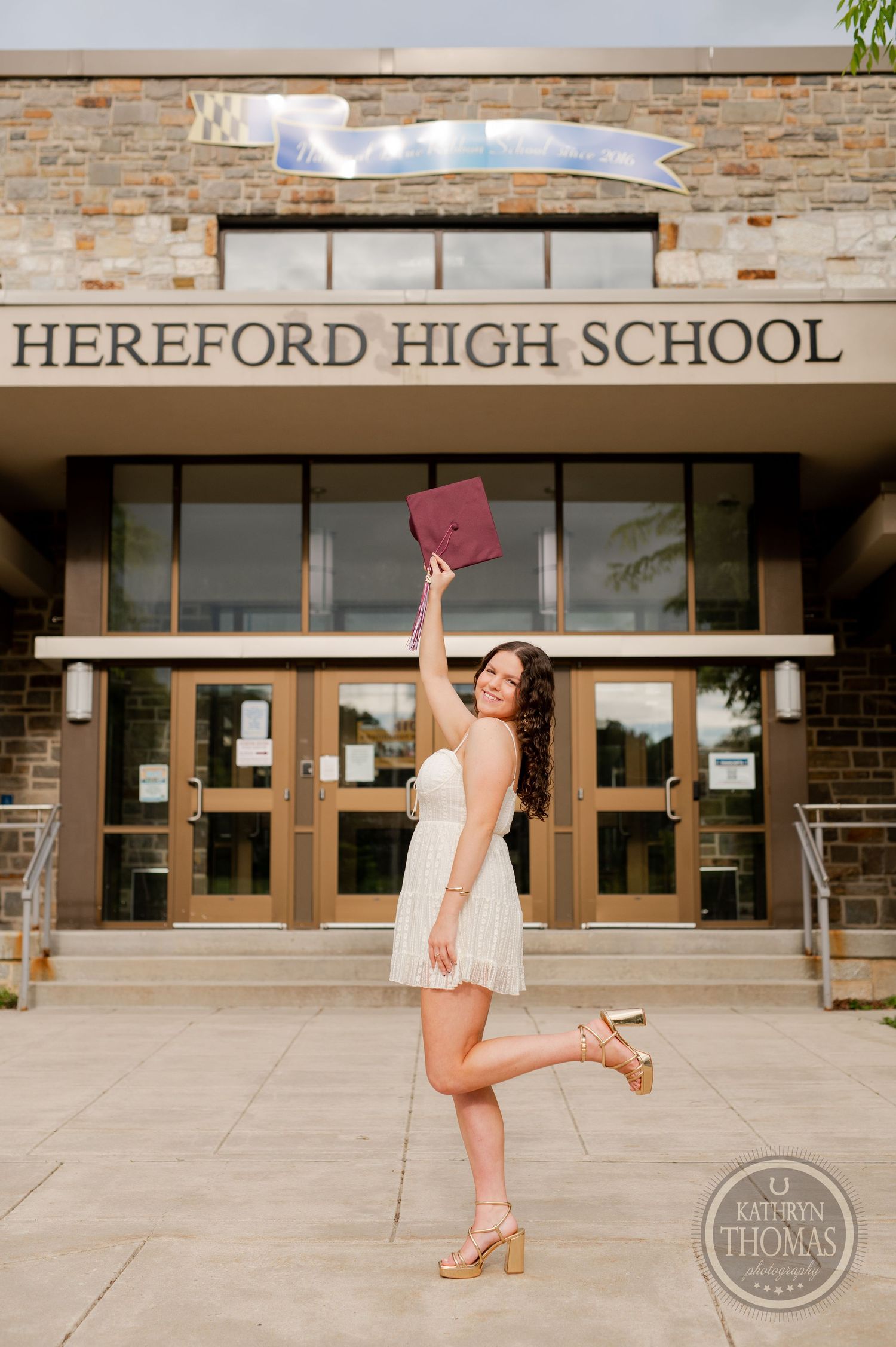 Graduate jumps joyfully while holding maroon cap in front of high school building.