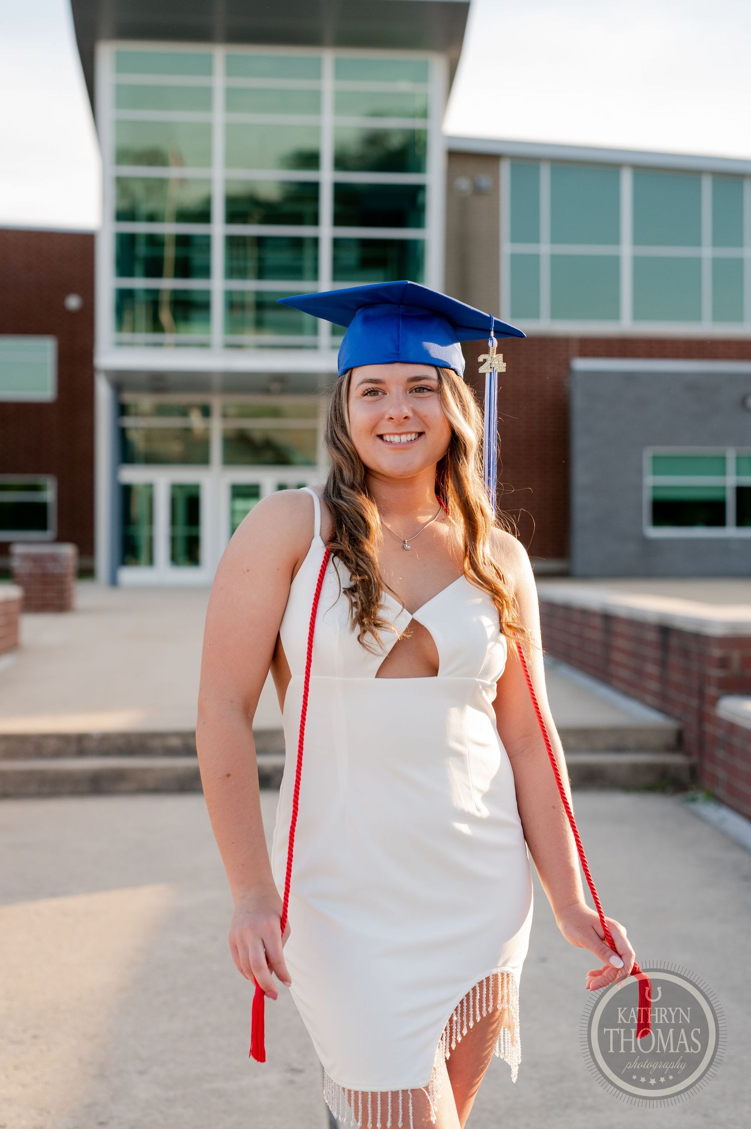 A graduate wearing a blue cap and white dress poses in front of a school building entrance.