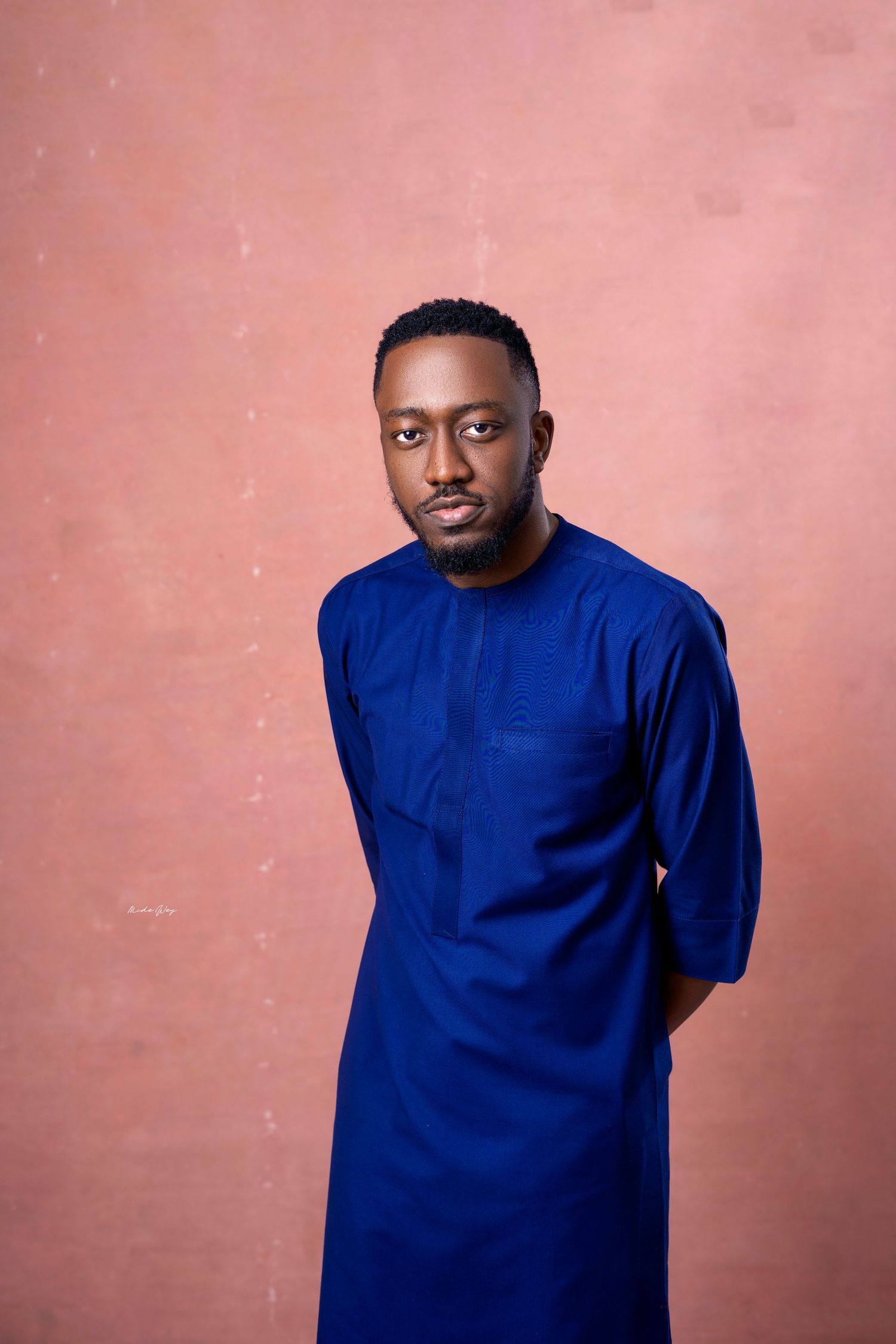 Person wearing traditional blue Nigerian agbada poses against coral colored background.