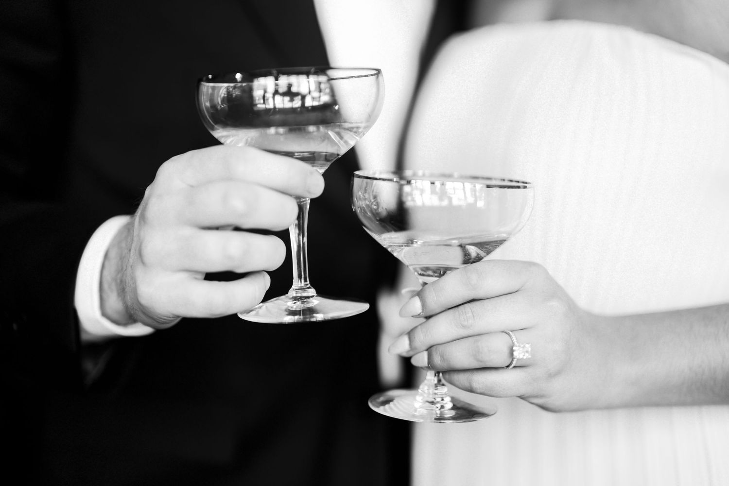 Black and white photo of two hands clinking champagne glasses in elegant stemware during a formal celebration.