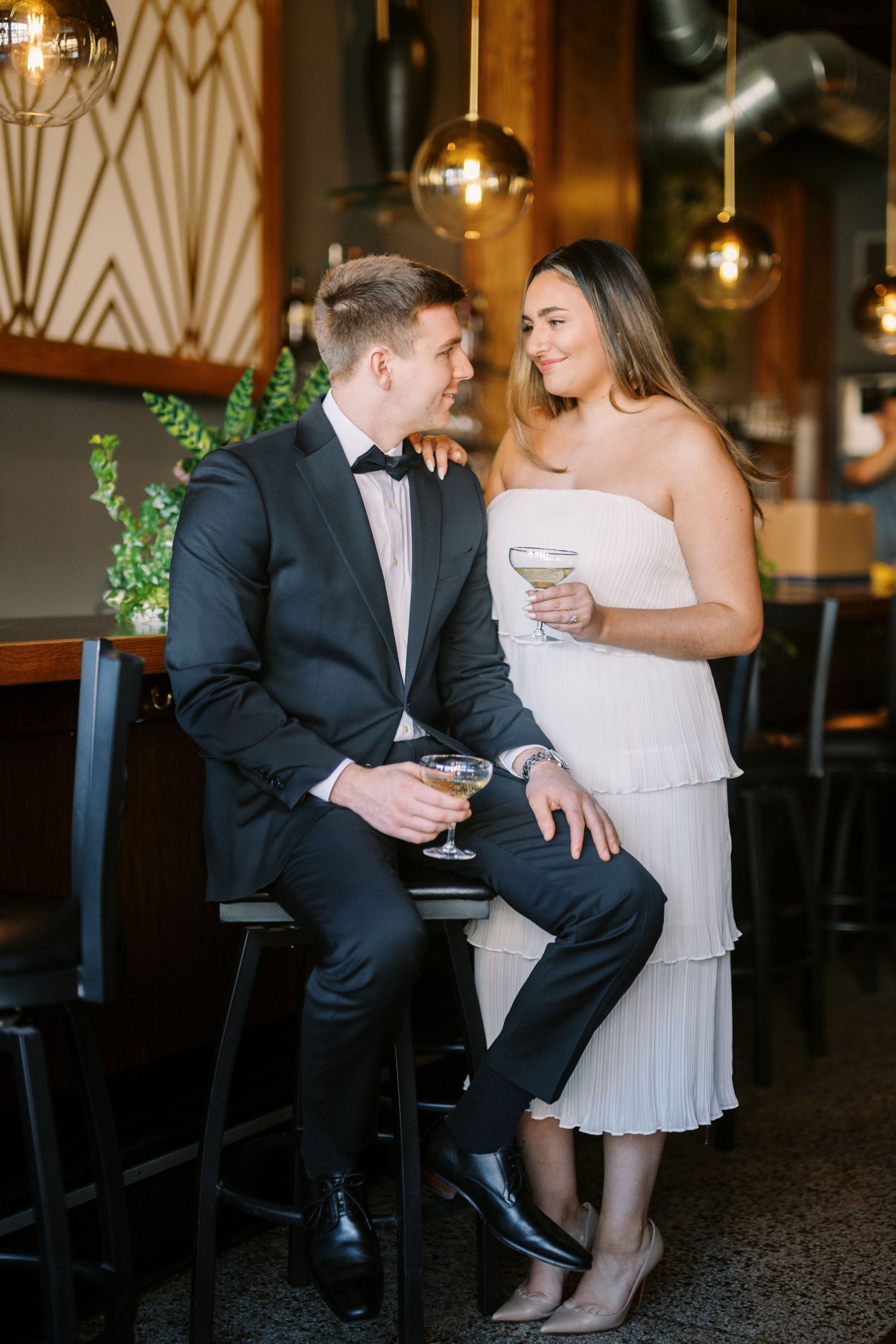 Couple in elegant attire sharing drinks at an upscale bar with art deco decor and hanging pendant lights.