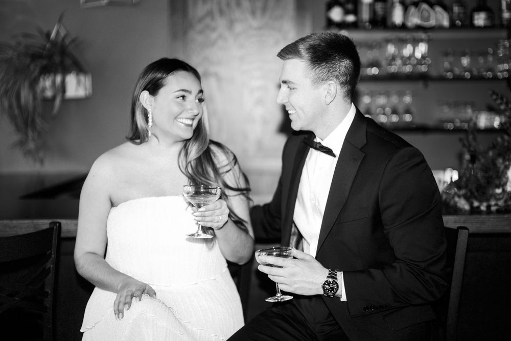 A black and white photo of a couple in formal attire engaged in conversation at a bar with wine glasses.