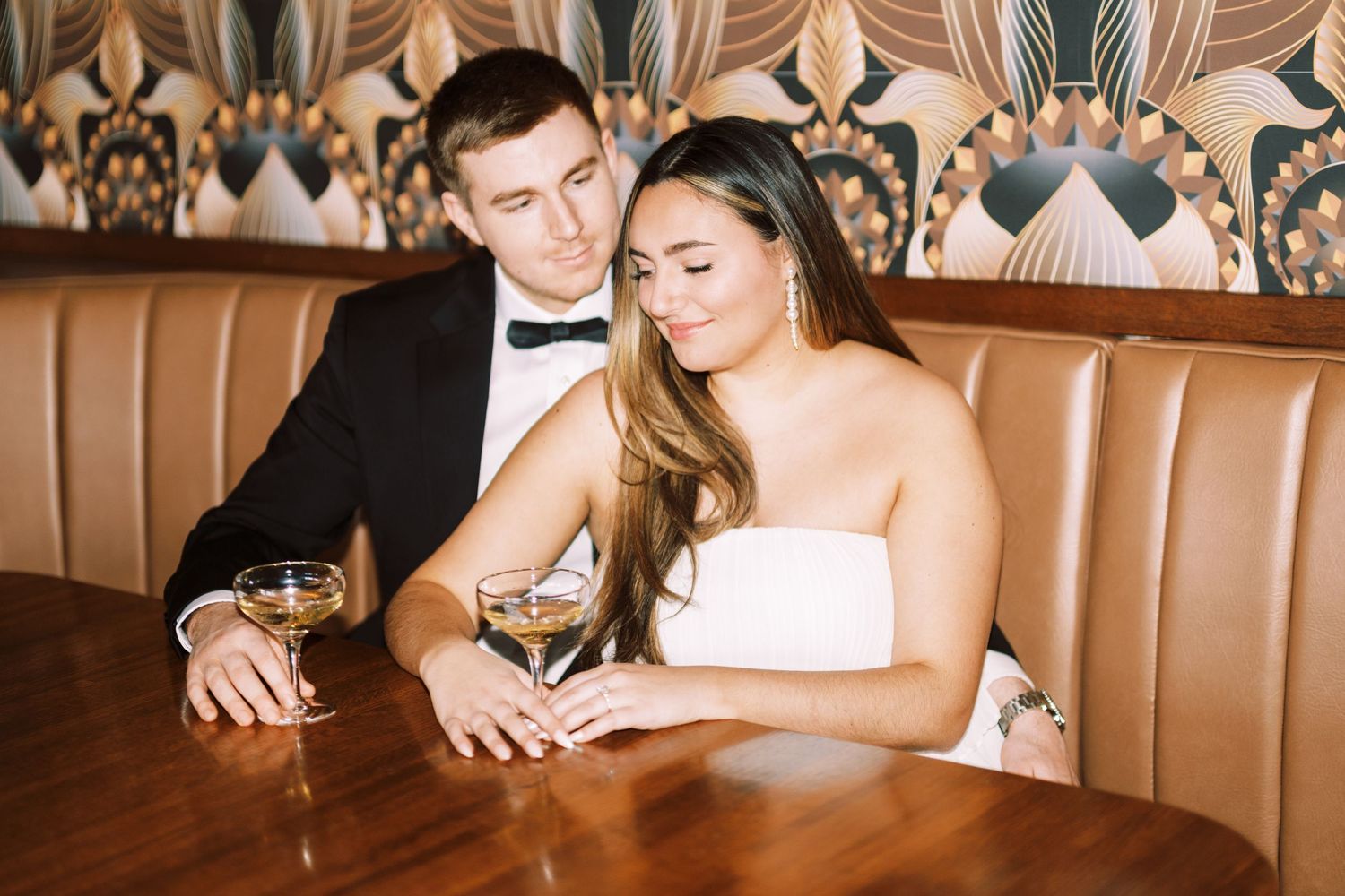 Elegantly dressed couple sharing an intimate moment at a brown leather booth with decorative wallpaper behind them.