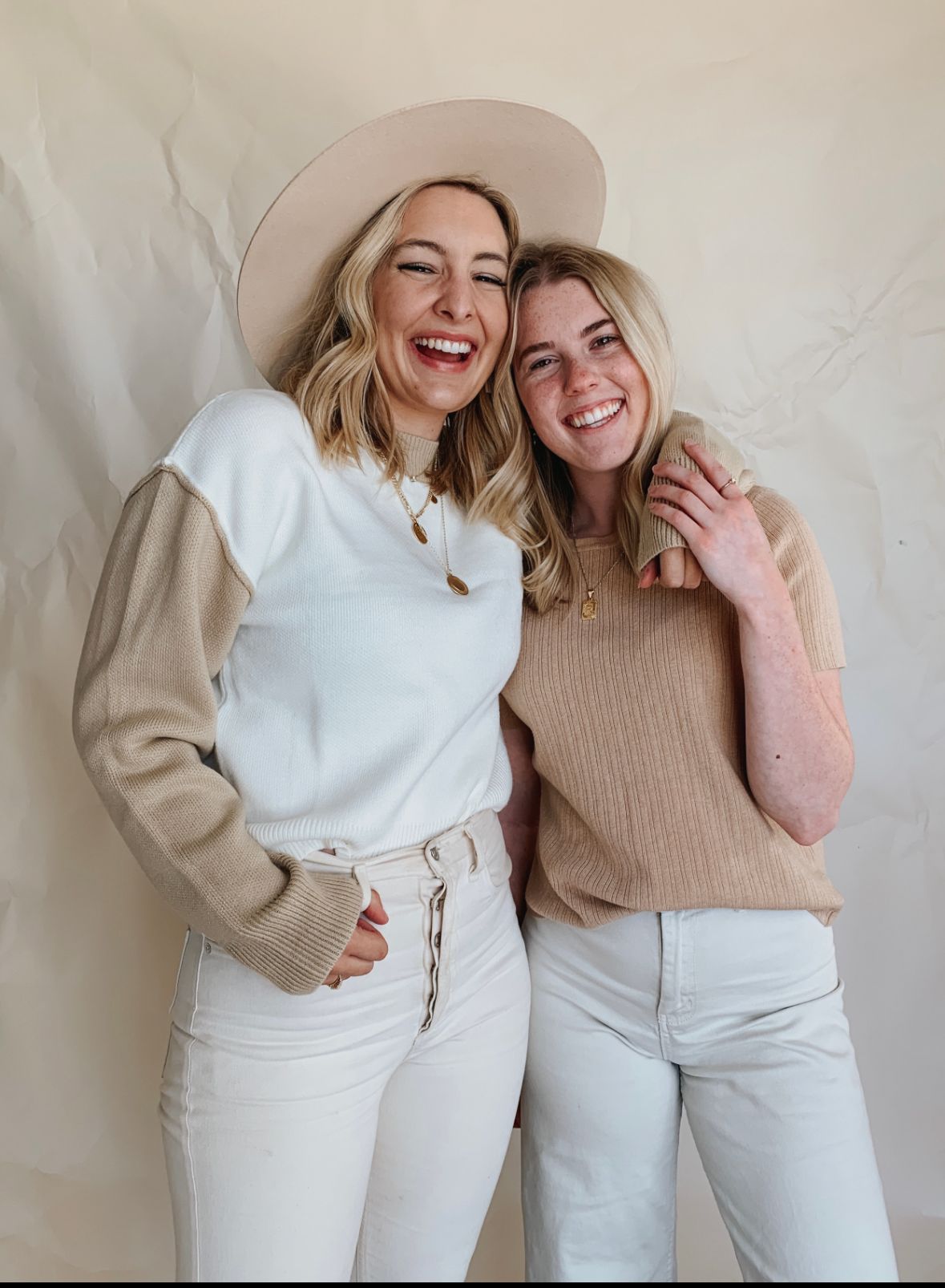 Two friends in neutral-toned outfits share a candid laughing moment against a white backdrop.