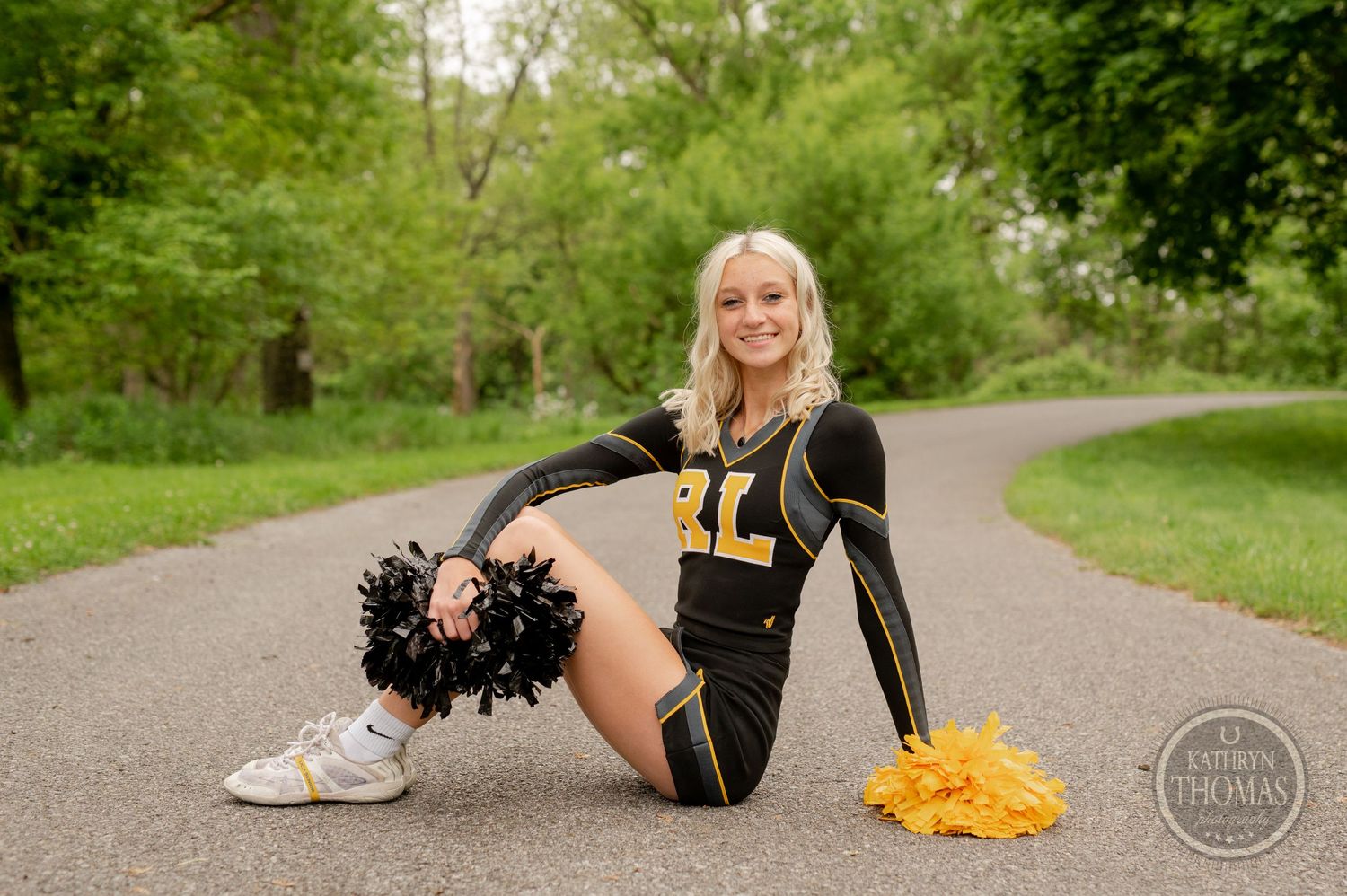 Performer in black uniform poses with black and gold pom poms on park pathway.