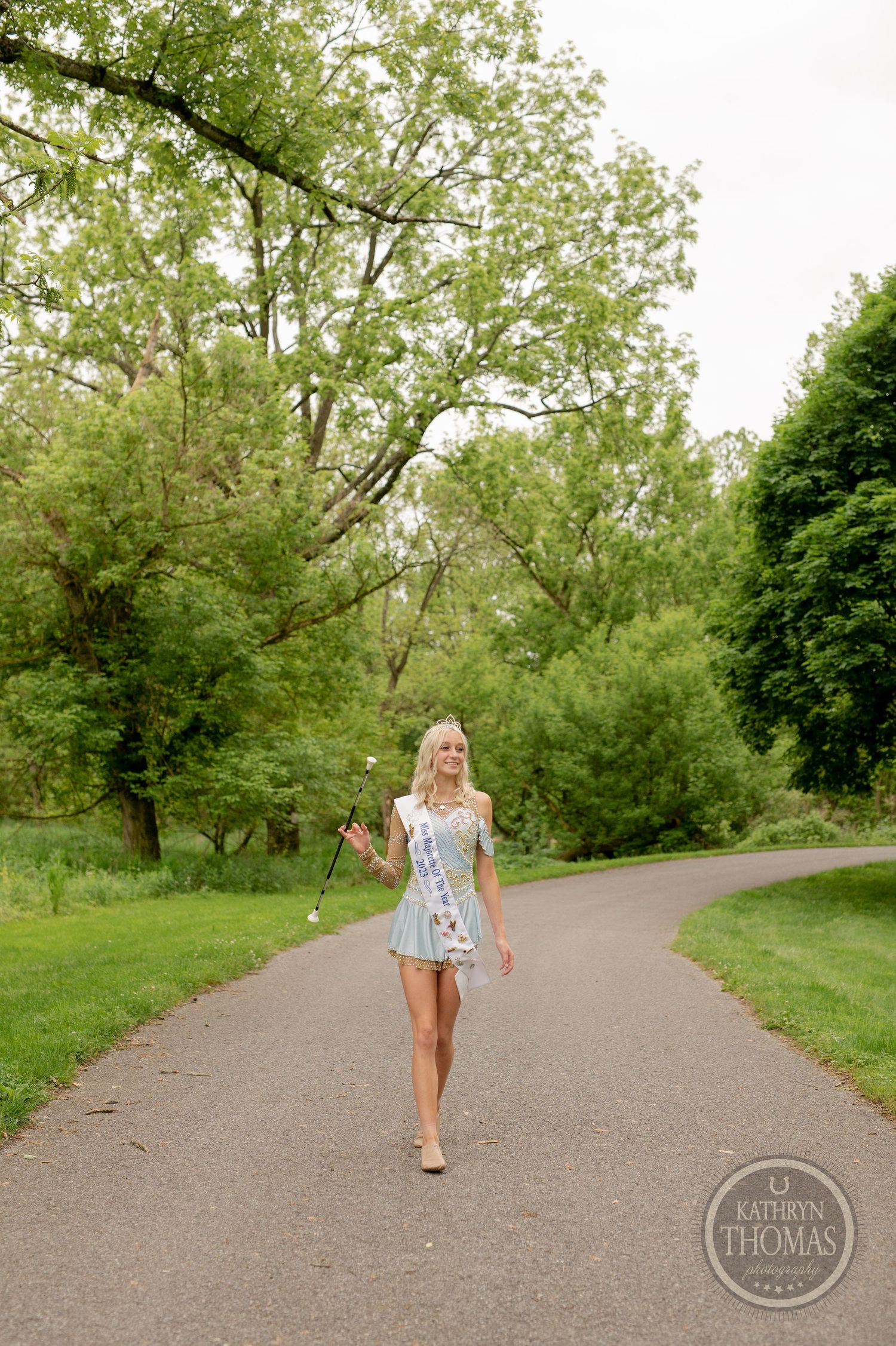 Pageant contestant in white dress walks along park path surrounded by spring greenery.