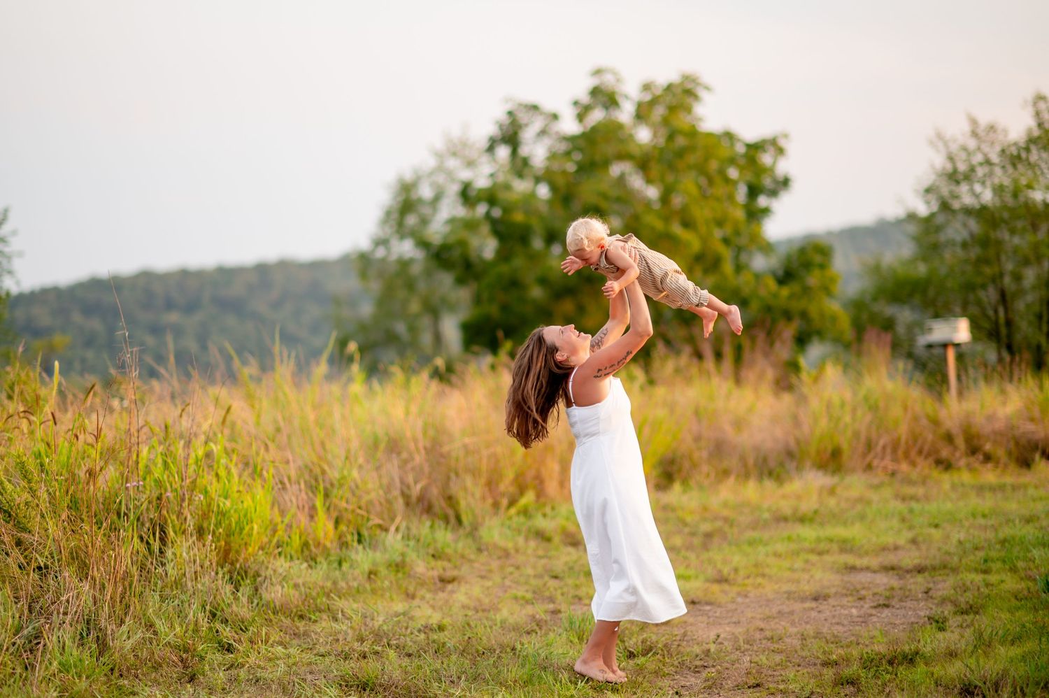 A parent wearing a white dress lifts a child joyfully in a golden summer meadow with rolling hills in the background.
