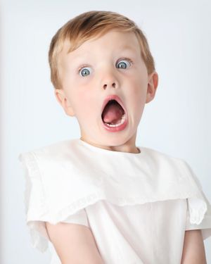 An excited child in white clothing shows surprise with wide eyes and open mouth.