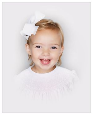 A delighted toddler with blonde hair wearing white dress and bow laughs against light backdrop.