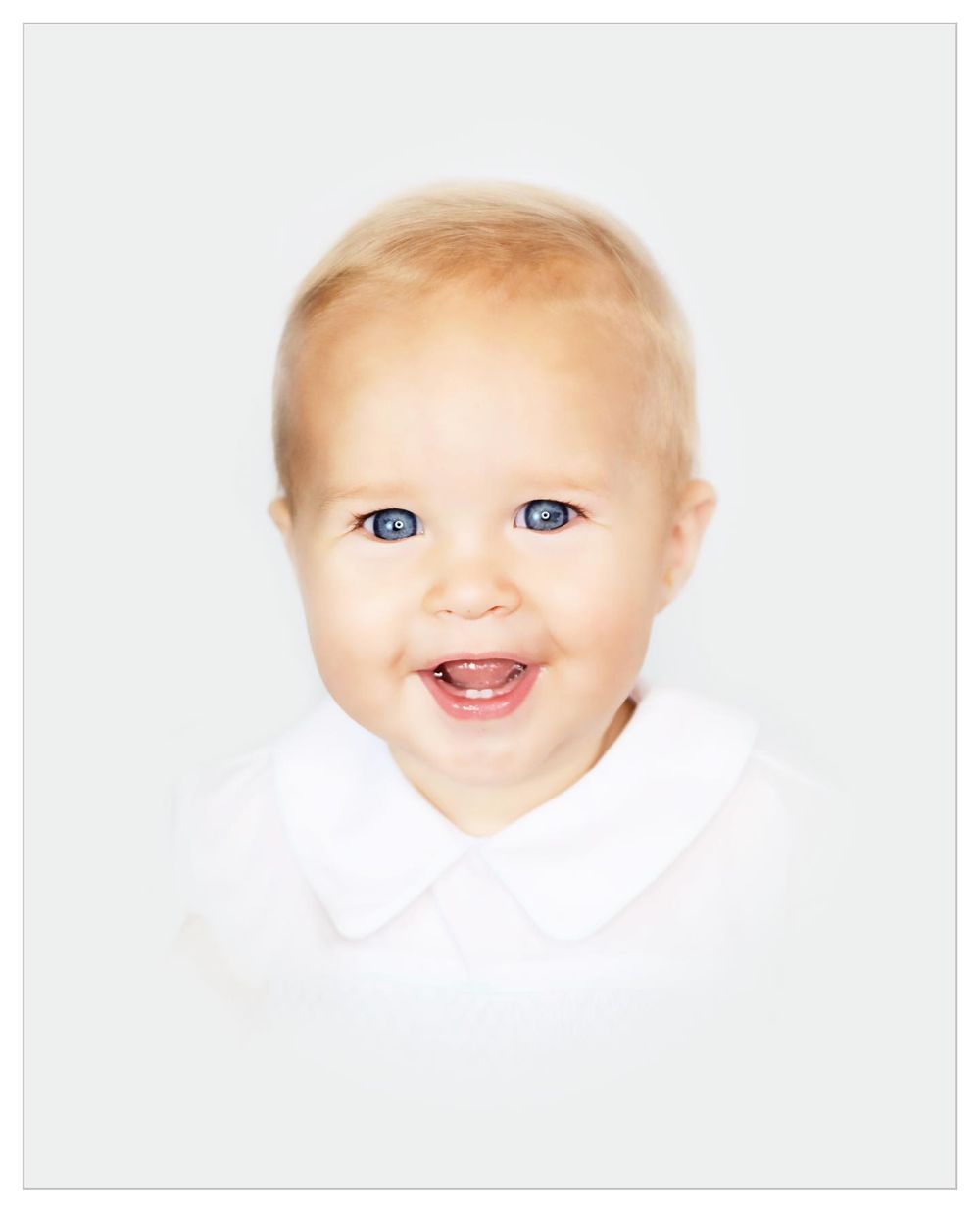 Studio portrait of a smiling baby in white clothing against neutral backdrop.