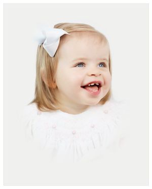 A joyful toddler wearing a white dress and bow laughs while looking up against a light background.