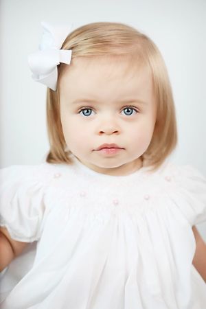 A young child in a white dress and bow poses against a neutral background with bright blue eyes.