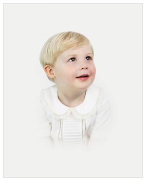 Professional portrait of a small child in white clothing looking up with a gentle expression against a light background.