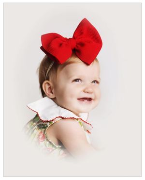 A cheerful toddler wearing a large red bow and floral outfit poses against a light background.