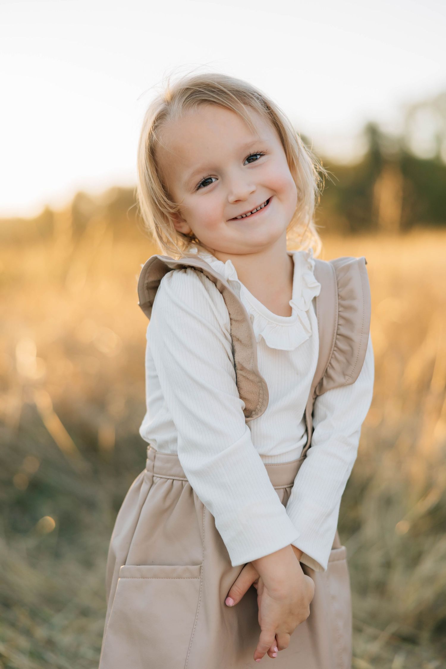 Adorable blonde toddler girl smiling in a ruffled dress in a golden sunlit field.