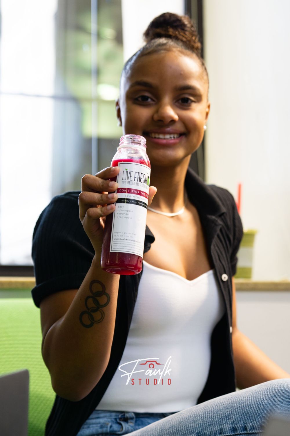 A person in a black jacket and white top holds a pink beverage bottle while relaxing on a couch.