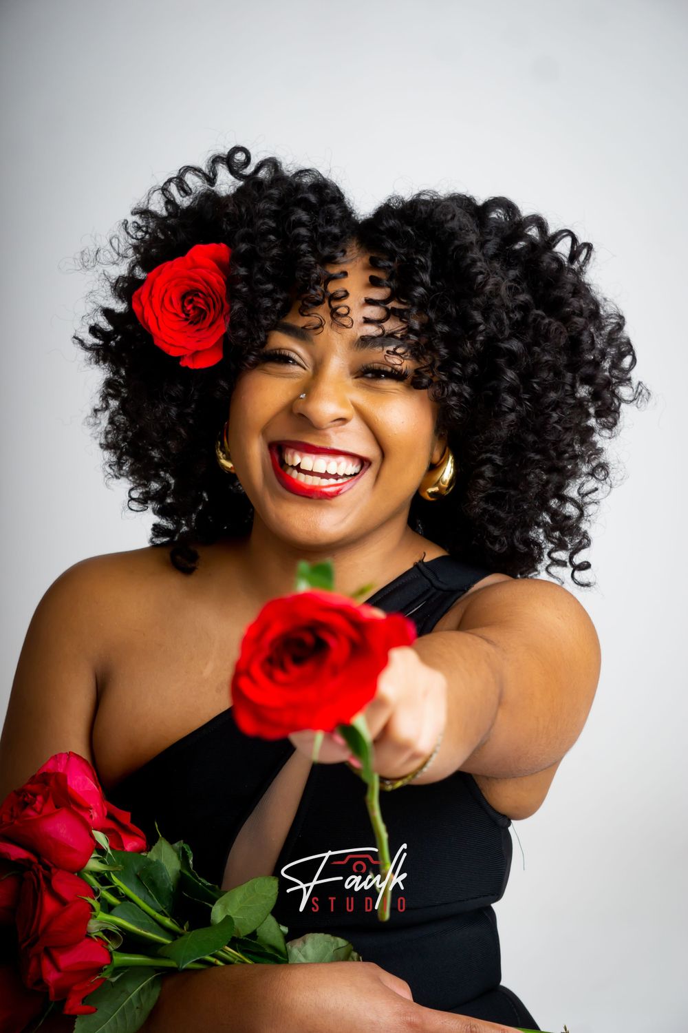 A woman in a black top poses with vibrant red roses against a light background, showcasing her natural curly hair.