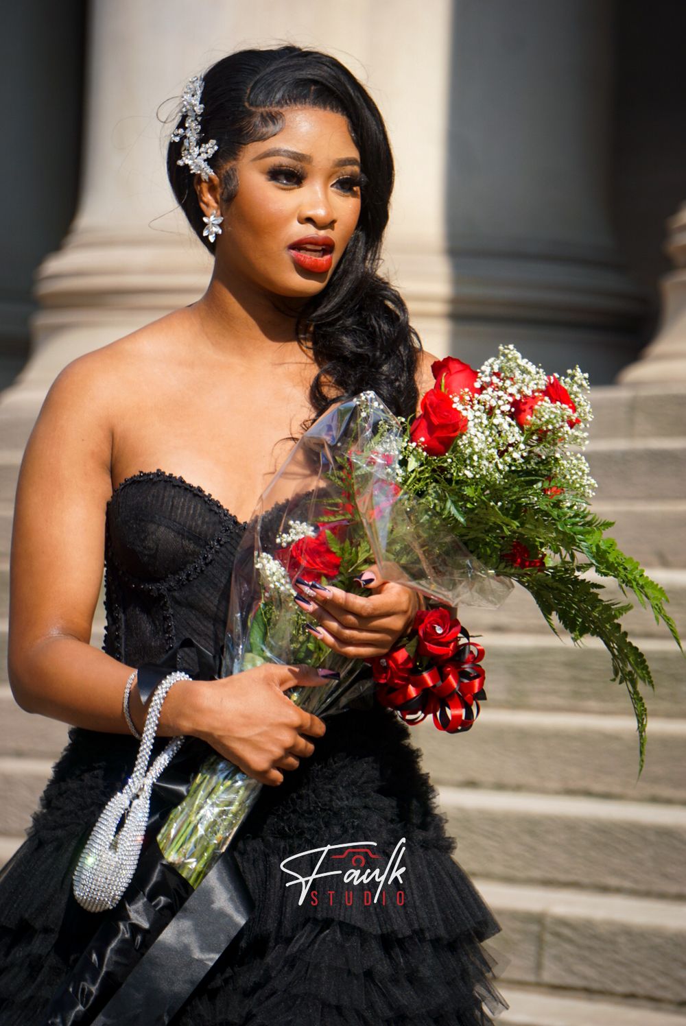 A person in a black strapless gown holds red roses and baby's breath bouquet while posing on stone steps.