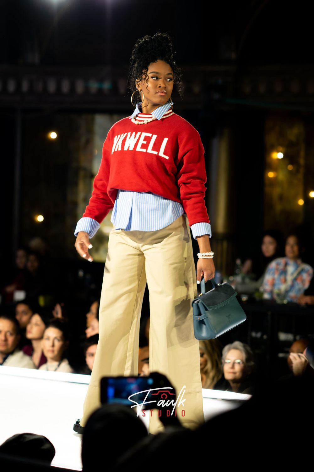 Model walks runway in red sweater and khaki pants during fashion show with audience in dark background.