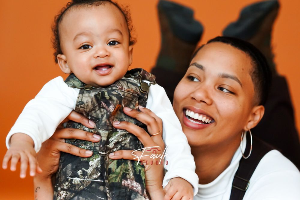 A joyful moment captured against an orange backdrop with a baby wearing camouflage overalls and white shirt.