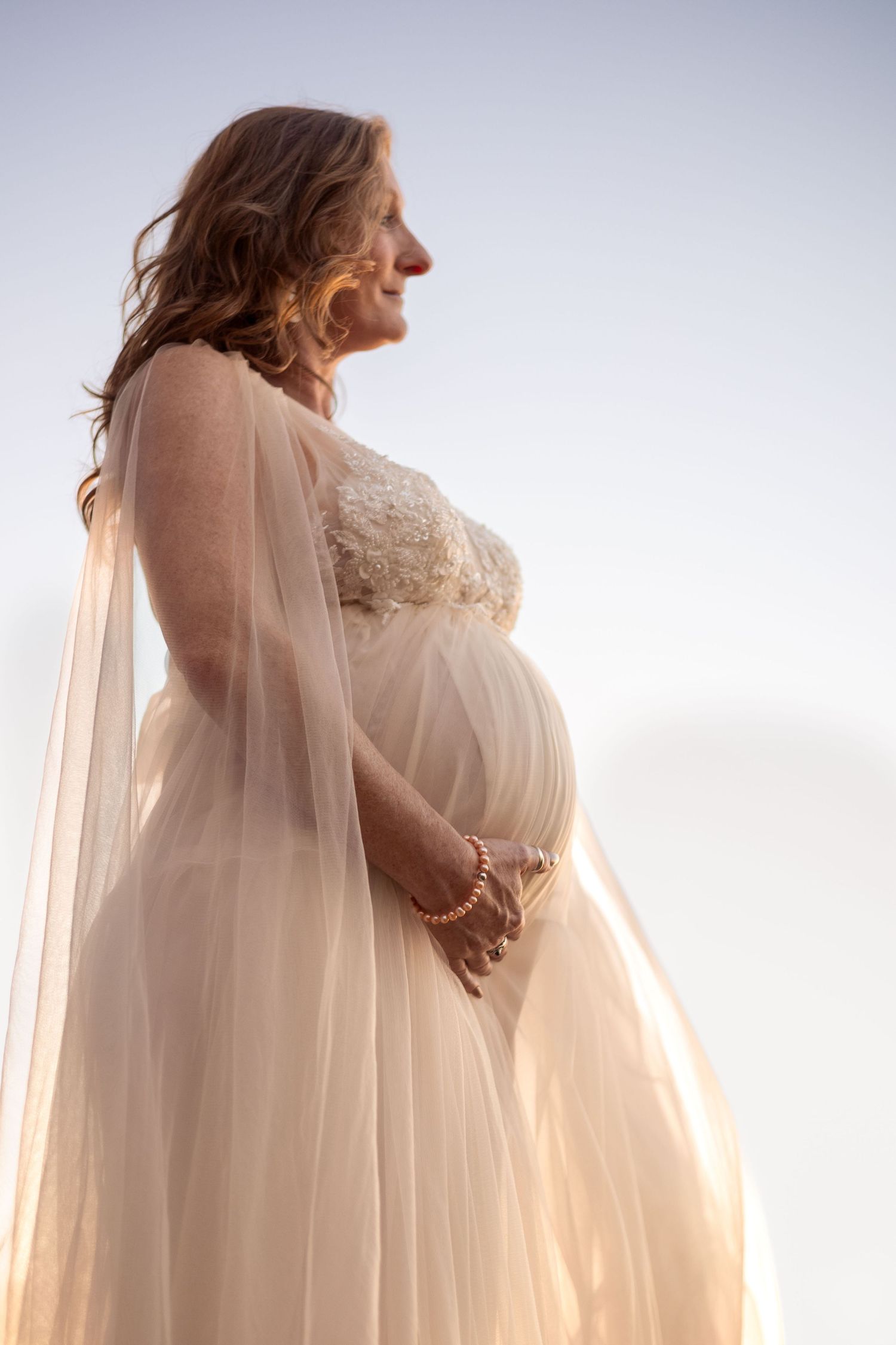 A pregnant woman is photographed from a low angle and looks off to the distance while cradling her belly.