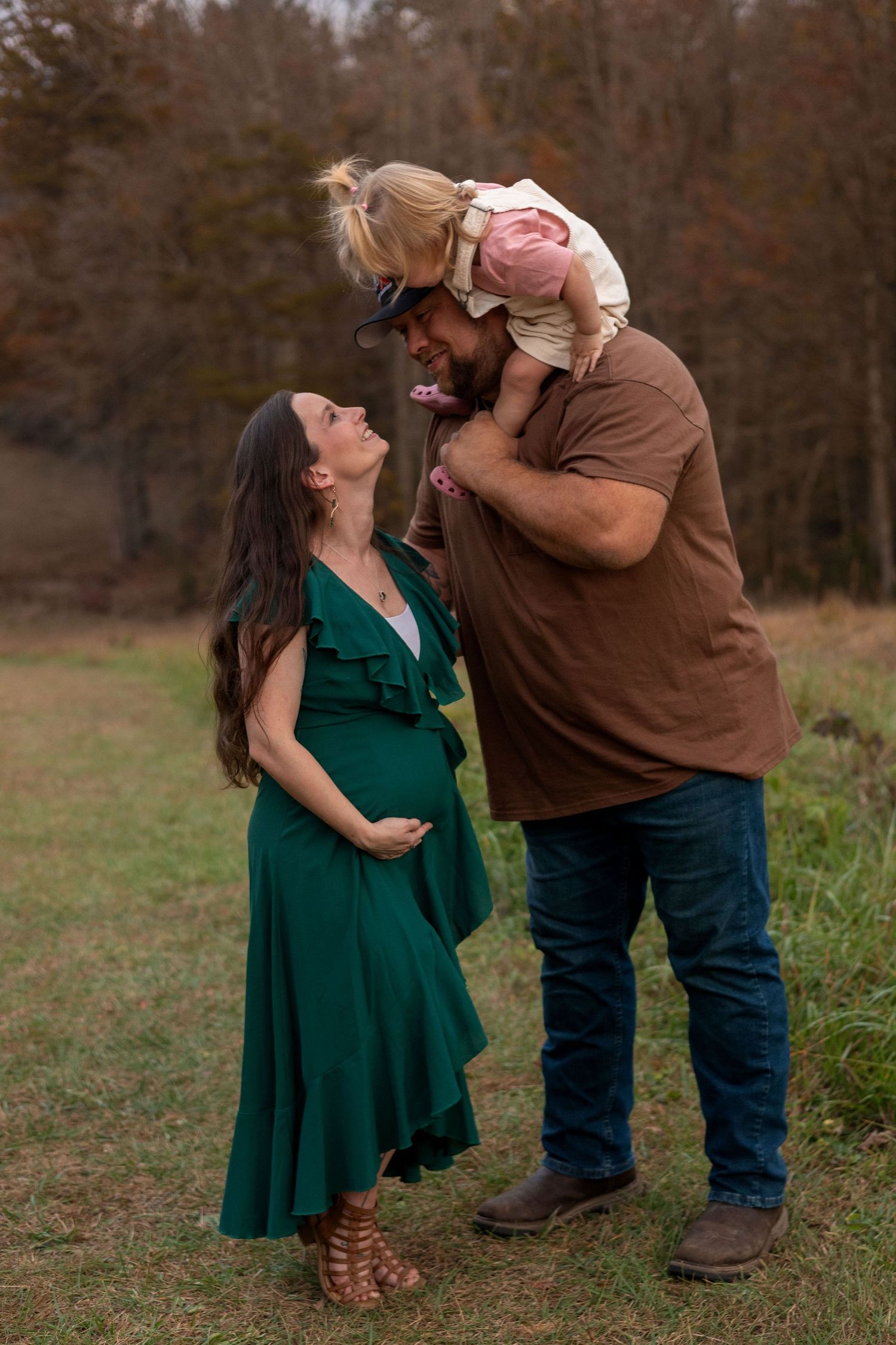 A very tall dad holds his toddler on his shoulders while she leans down to kiss her mom.
