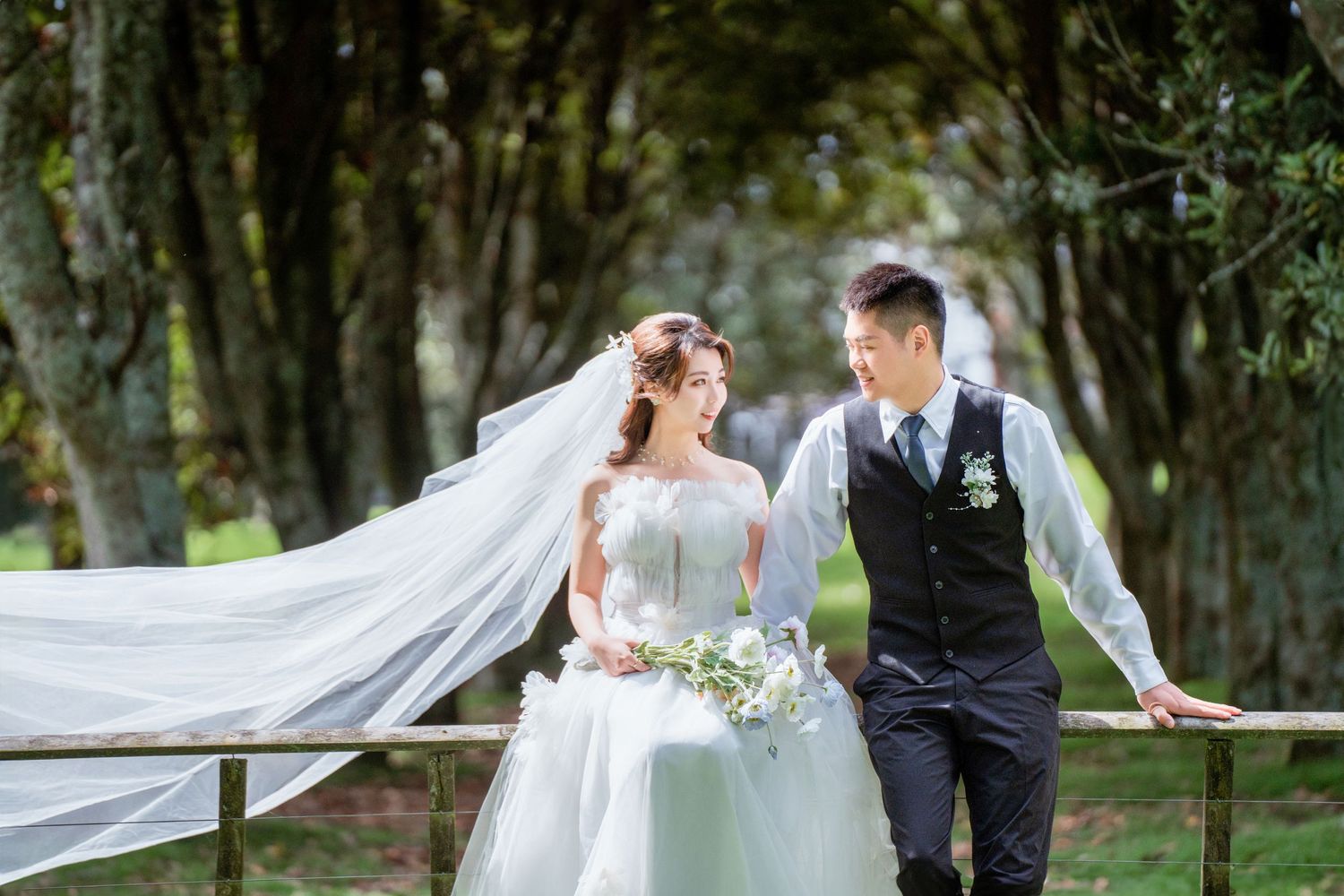 Bride and groom walking by a wooden fence in a lush green Auckland woodland setting for outdoor pre-wedding photography.