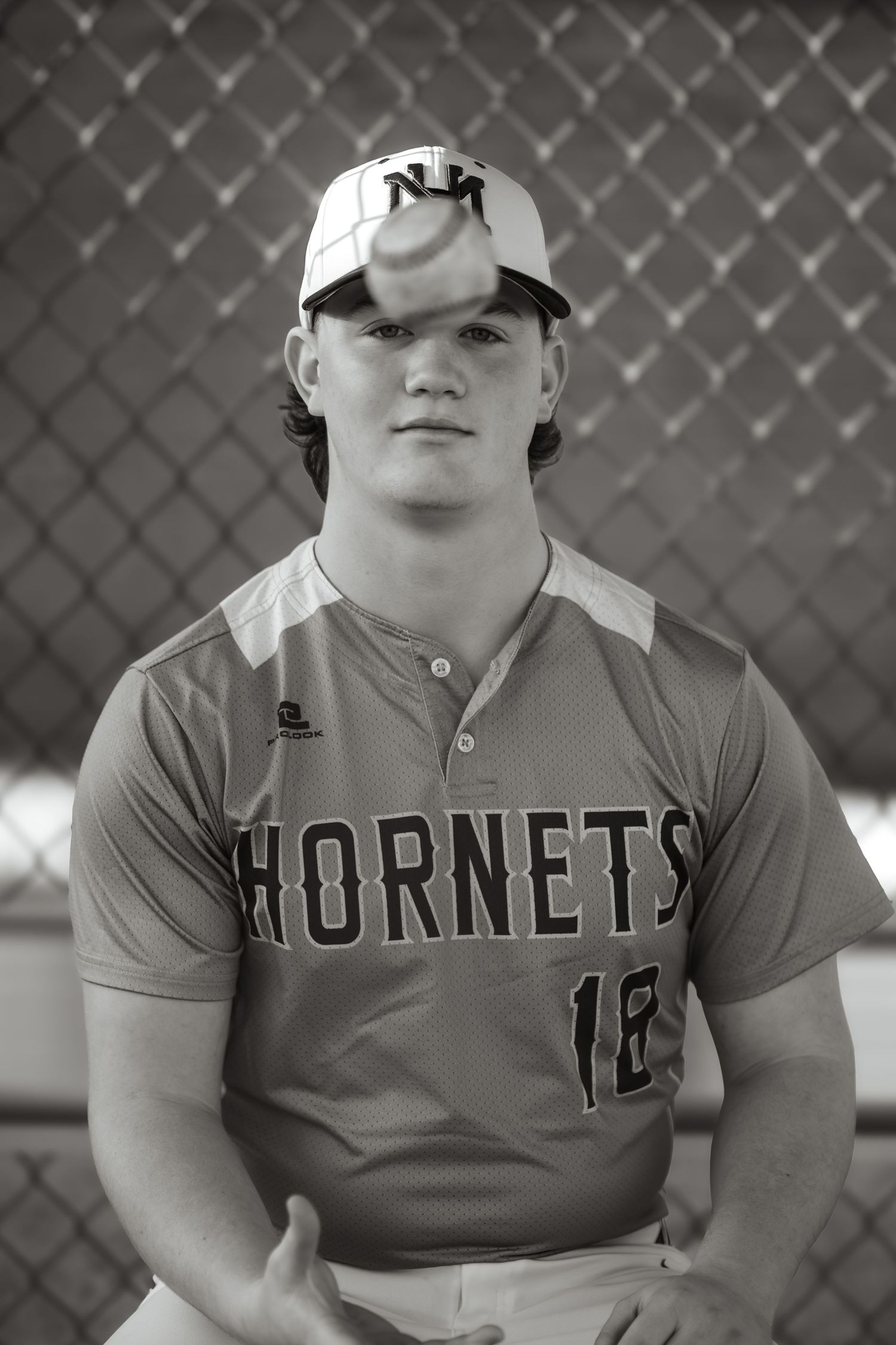 Baseball player in Hornets uniform photographed against chain link fence in black and white, celebrating his senior year of high school.