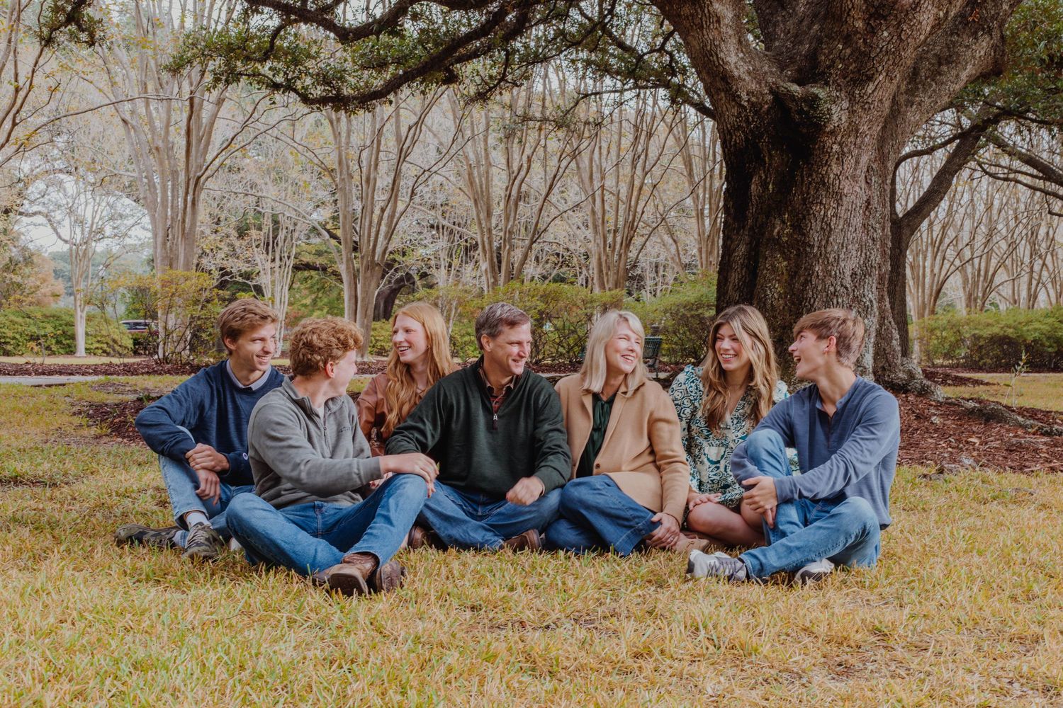 Friends sitting in a row on grass laughing together under oak trees.