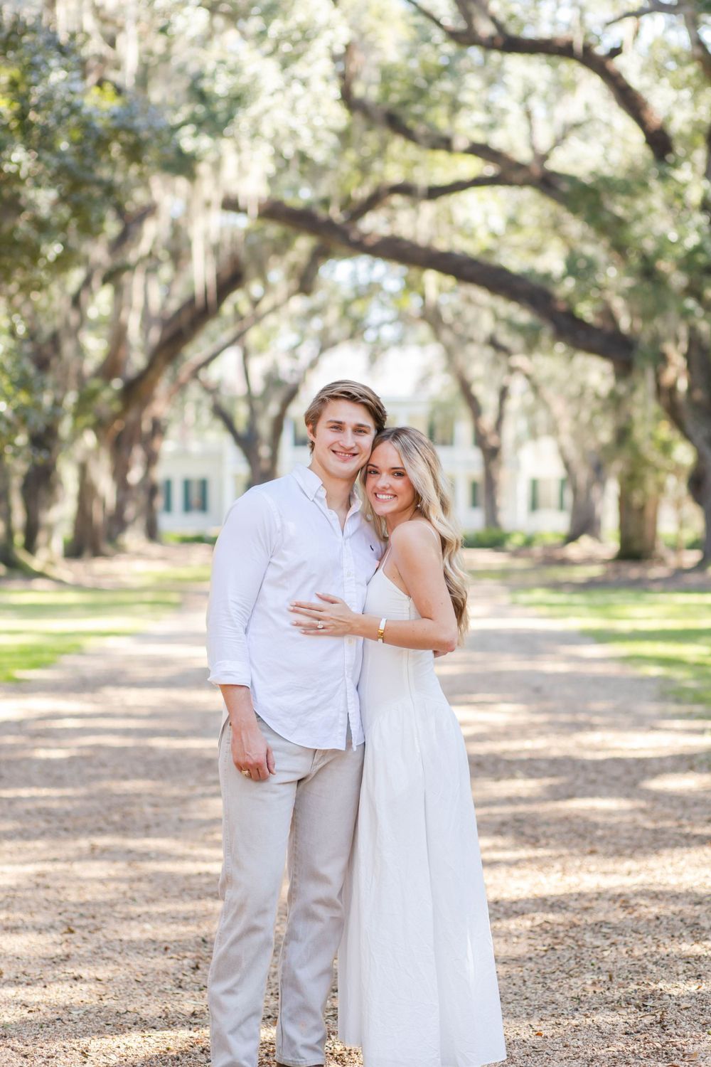 Couple in white and khaki poses on tree-lined path with Spanish moss canopy.