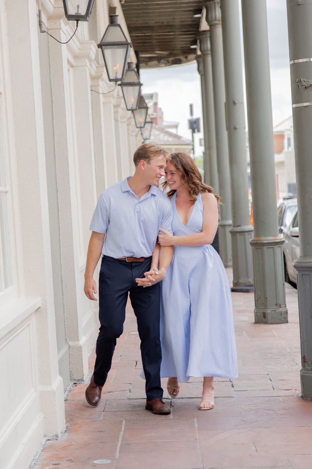 A couple in coordinating blue outfits strolls and laughs together under a covered walkway with gas lanterns.