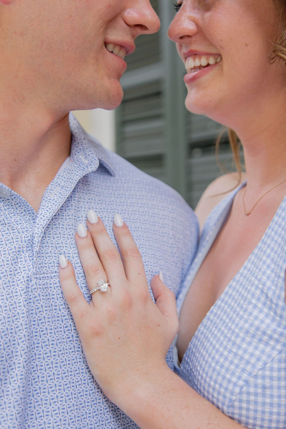 Close-up of engagement ring shown as couple in light blue outfits embrace and smile at each other.