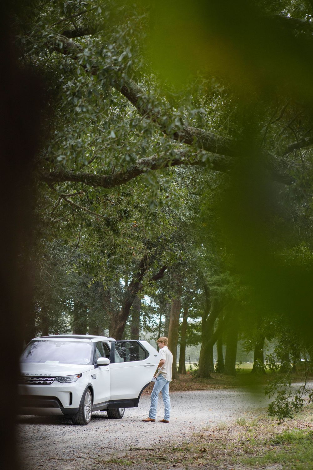 Person standing next to white car on tree-lined dirt path.
