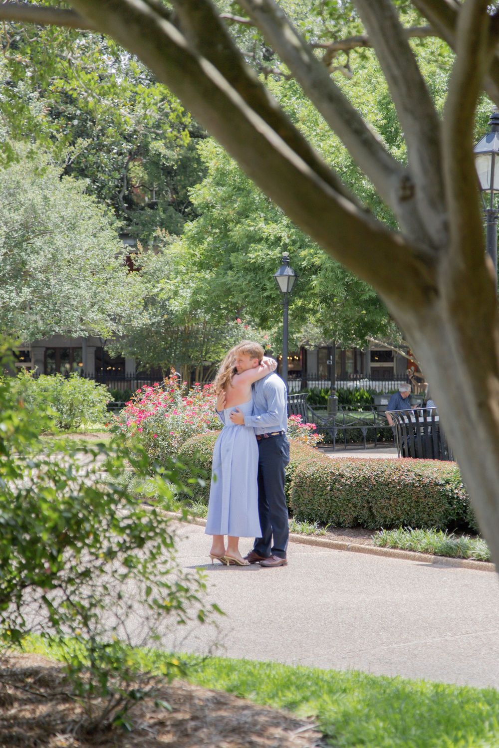 A romantic moment between two people embracing on a garden path with lush greenery and flowers surrounding them.