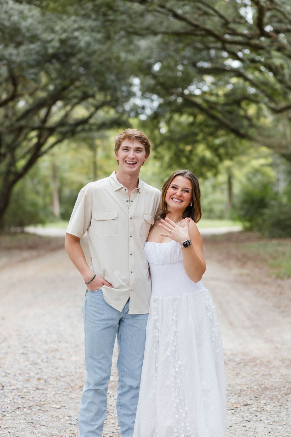 Newly engaged couple poses on tree-lined path showing ring.
