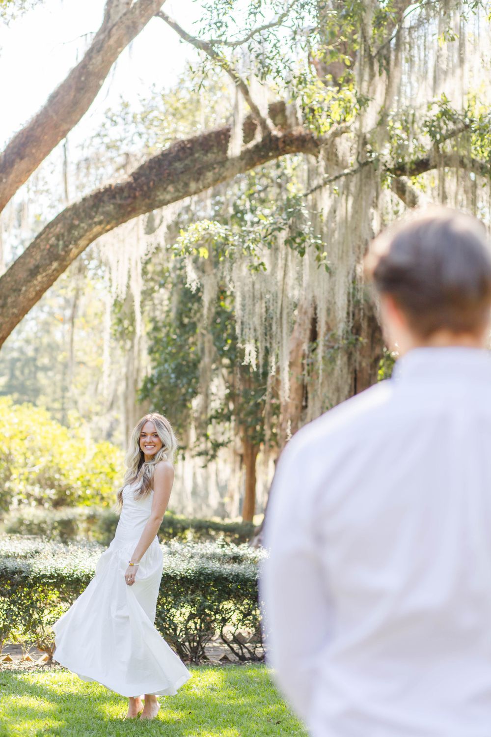 Playful bride twirls in white dress on lawn with Spanish moss trees overhead.