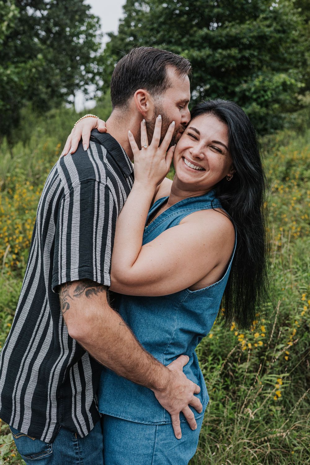 A joyful couple embraces outdoors, woman in denim overalls laughing as man kisses her cheek in a lush green field.