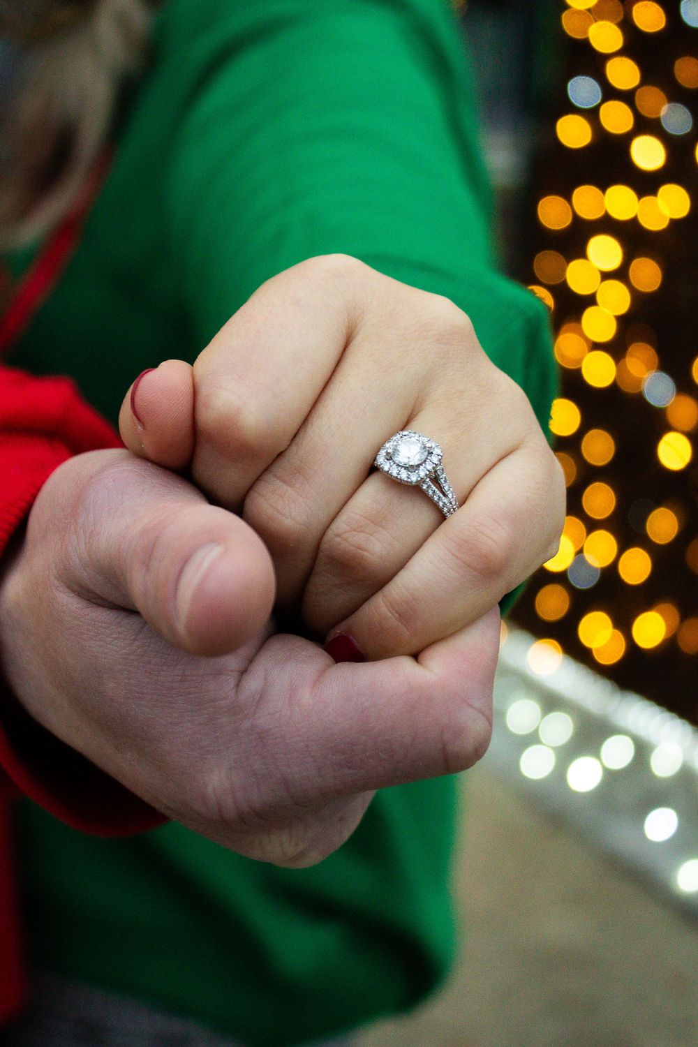 Diamond engagement ring shown on hand with festive holiday lights in background.