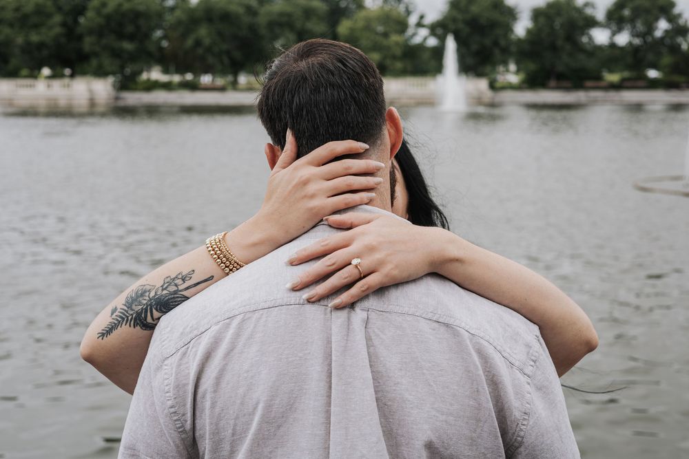 Couple embracing near a lake with a fountain, viewed from behind, man with tattoo wearing gray shirt.