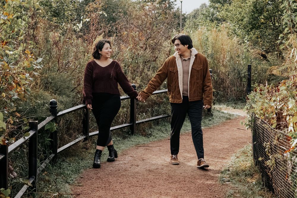 Two people walking and chatting on a scenic autumn path surrounded by fall foliage and a rustic railing.