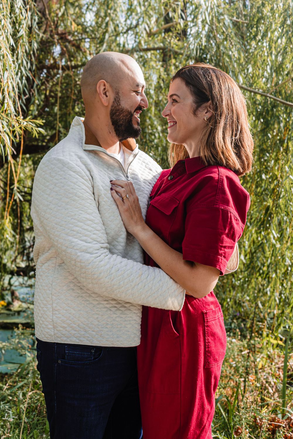 Couple embracing outdoors near willow trees, wearing burgundy and white casual attire.