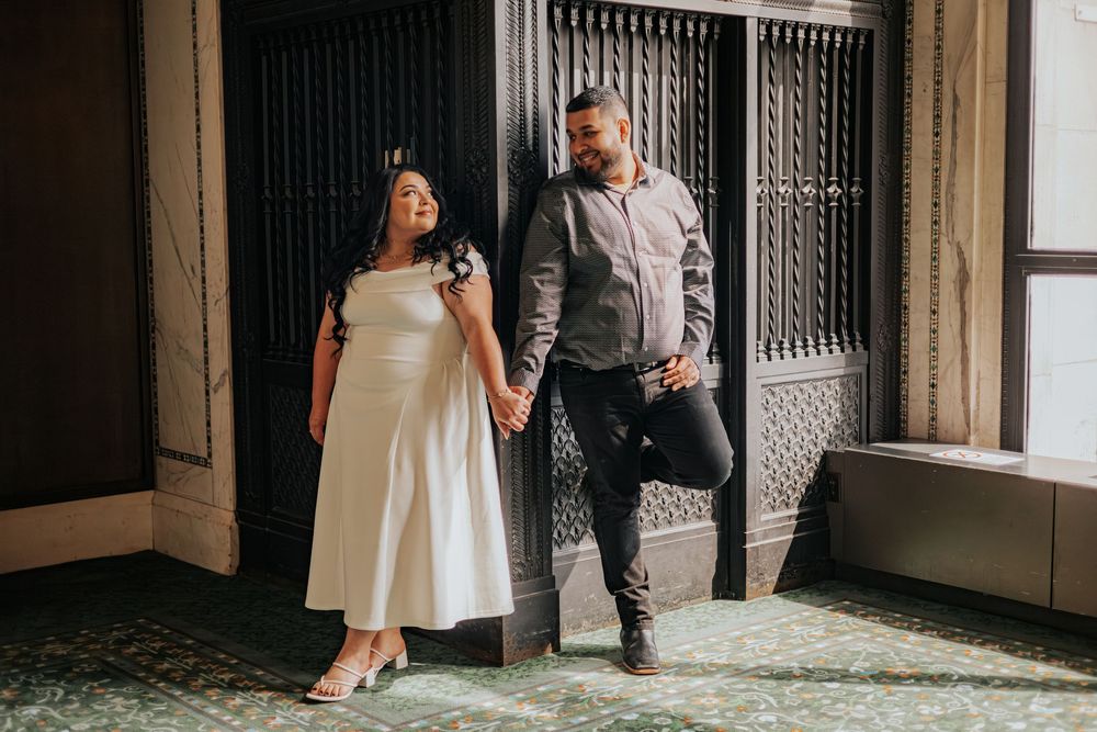 Couple holding hands and smiling in elegant indoor venue, woman in white dress and man in gray shirt.