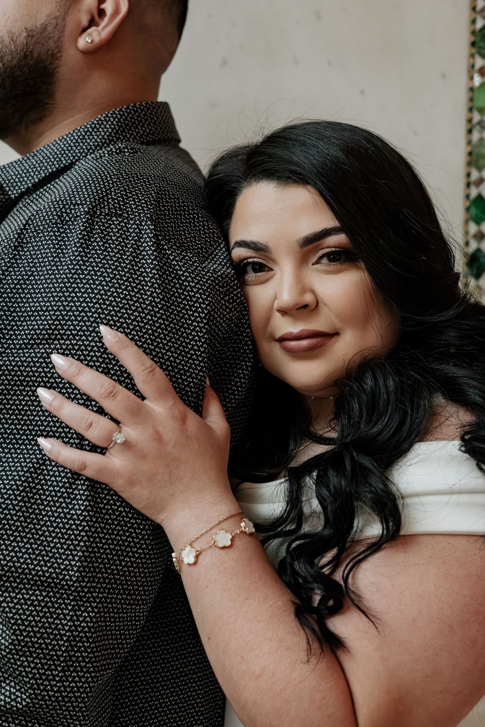 A woman with dark hair hugs a man in a black shirt, showcasing a floral bracelet and ring while smiling softly.
