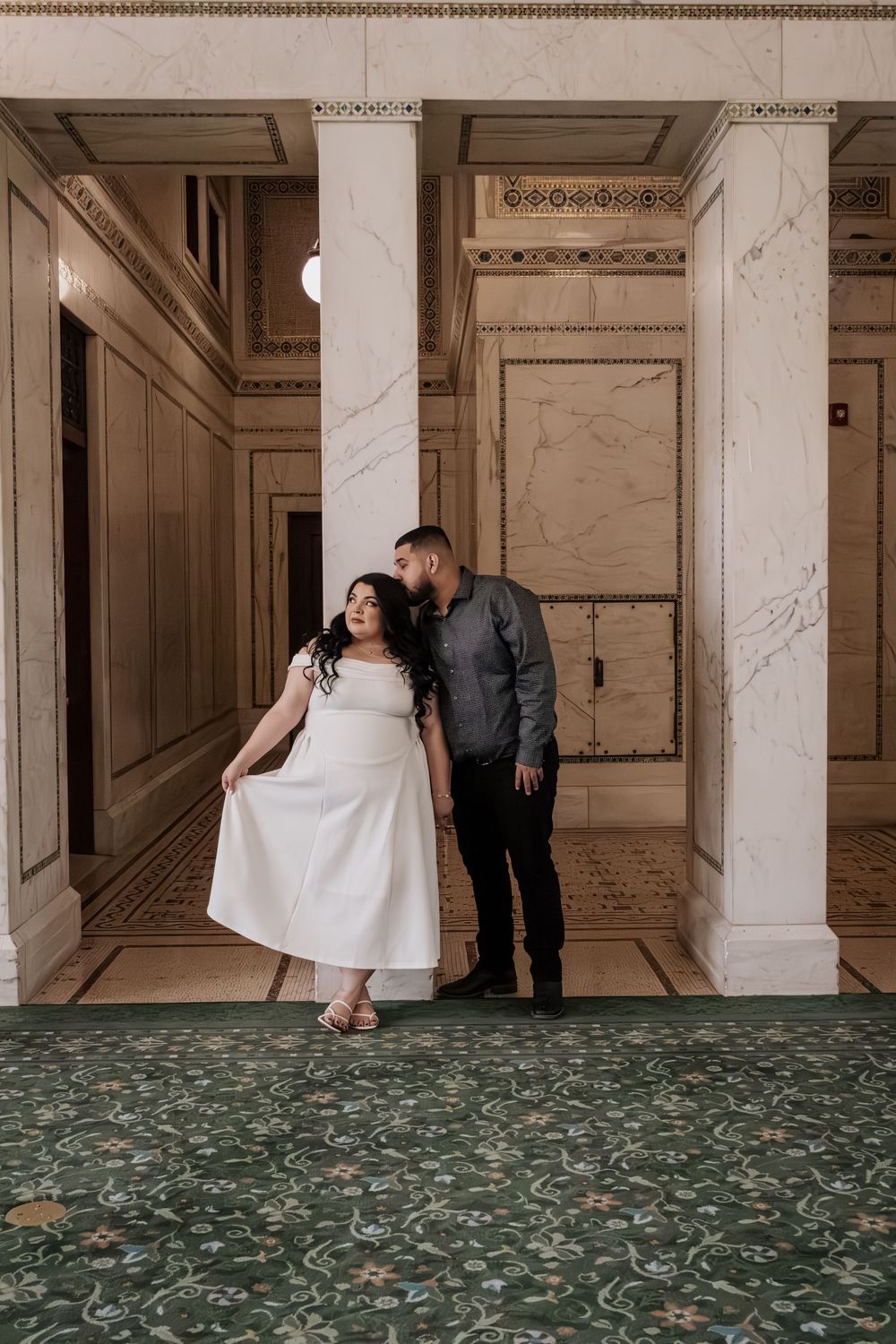 Couple in elegant marble hall, woman in white dress, man in gray shirt, standing on green floral carpet.