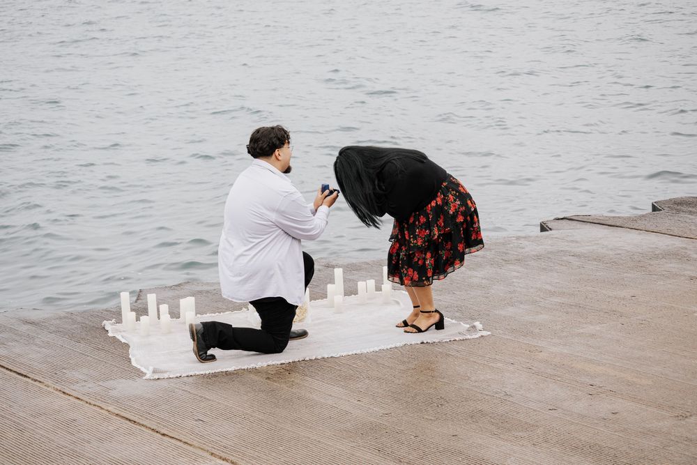 Man kneeling on dock proposing to woman near waterfront with candles on white blanket.